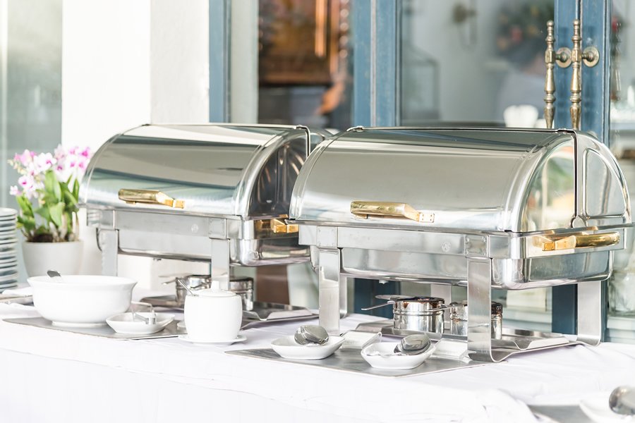 Two stainless steel chafing dishes with gold handles set on a white buffet table for a catered event.